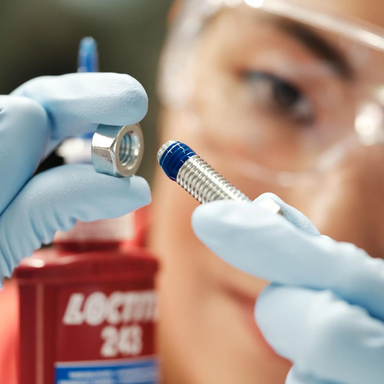 Close-up of a person with safety glassess. They have a bolt in one hand, and in the other hand a screw on which a blue LOCTITE threadlocker has been applied. The threadlocker bottle can be partially seen.