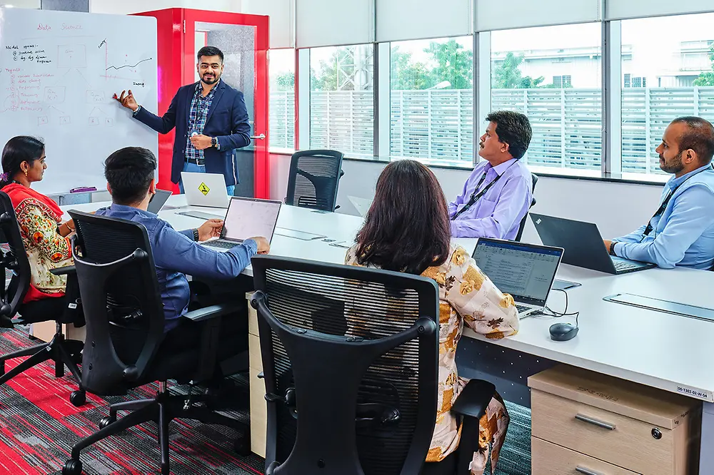 Five men and women are gathered at a conference table, listening to one team member explaining something on a whiteboard.