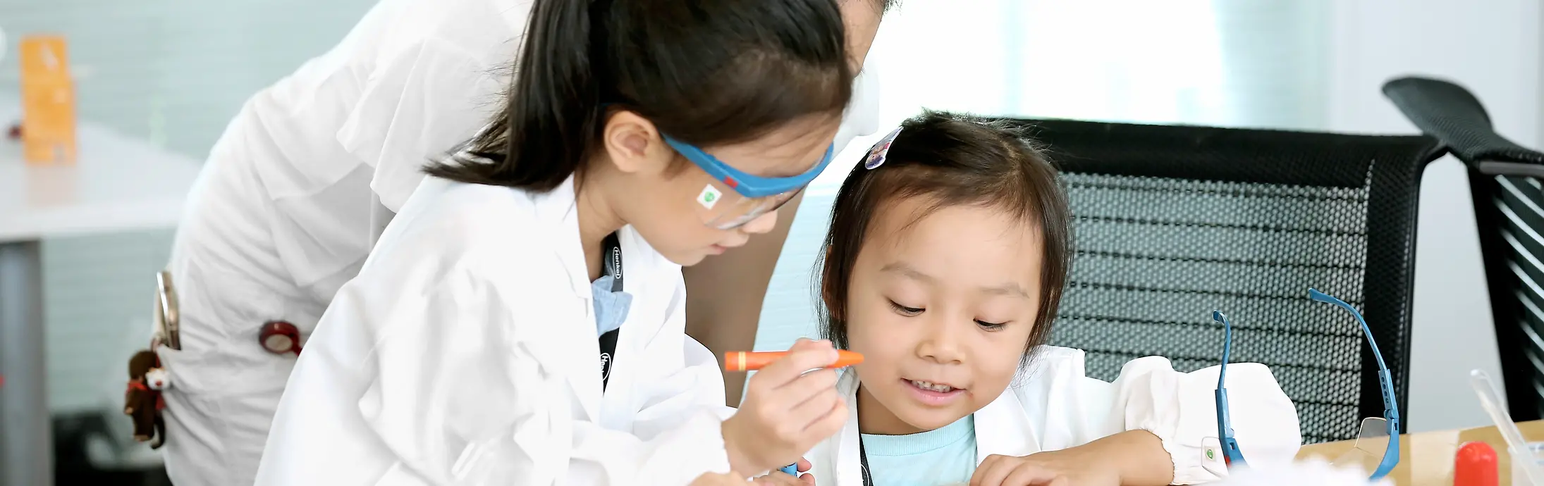 Two children and a woman in a research coat colour a picture at a table